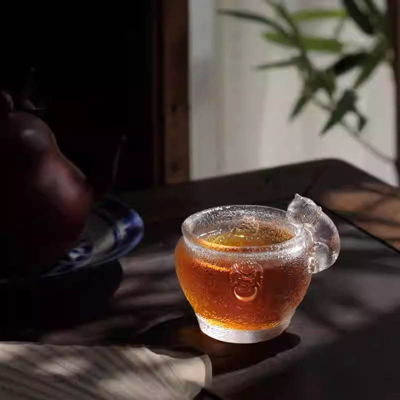 A handcrafted cat-themed glass teacup illuminated by natural sunlight, showcasing the sparkling ice-crackle texture and a warm tea glow on a wooden table.