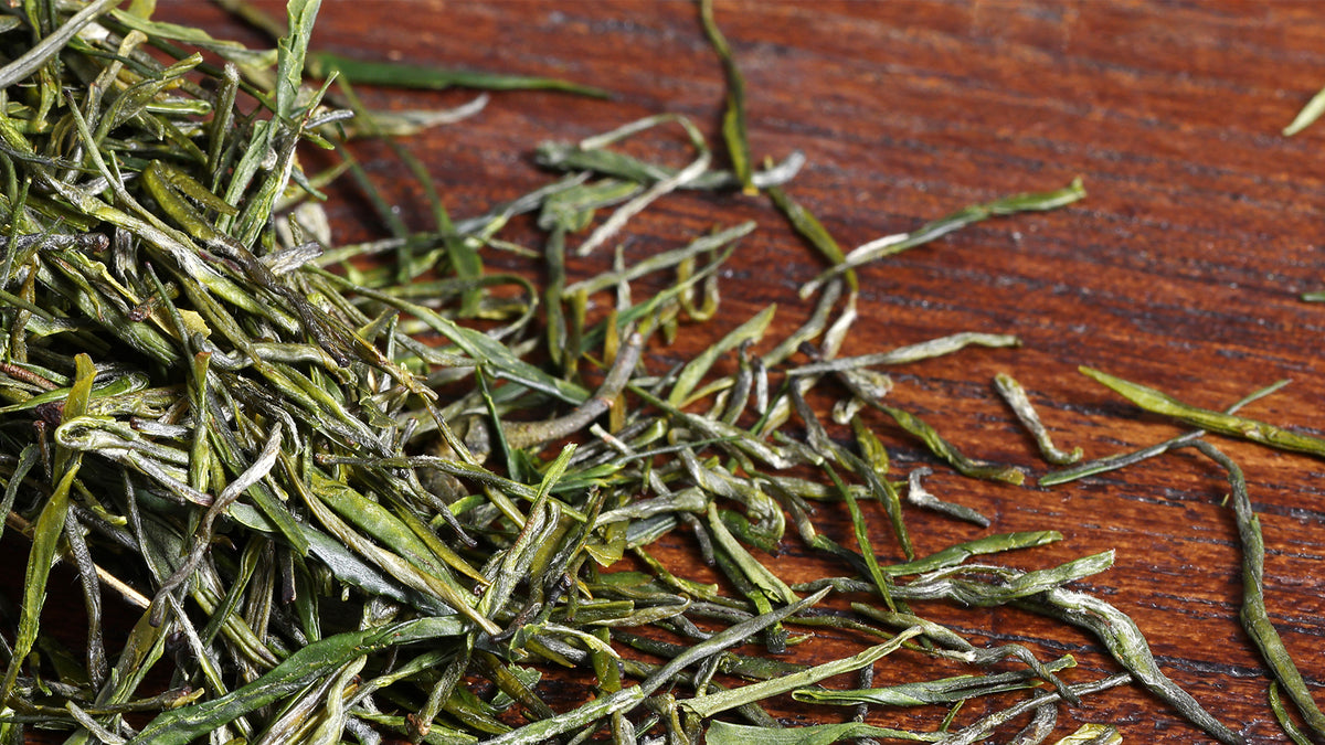 Close-up of loose-leaf Enshi Yulu green tea with slender needle-shaped leaves scattered on a dark wooden surface.
