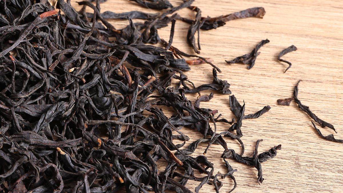 Close-up of loose-leaf wild black tea with dark twisted leaves scattered on a natural wooden surface.
