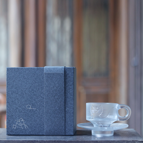 A sophisticated gift set featuring a frosted cat-engraved glass cup and saucer next to a premium dark grey gift box adorned with a minimalist mountain line drawing.