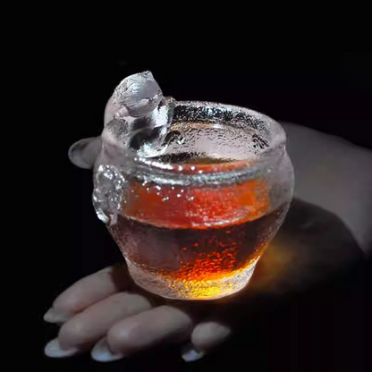 A handcrafted cat-themed glass teacup glowing with amber-colored tea, highlighting the playful kitty sculpture and sparkling hammered texture.