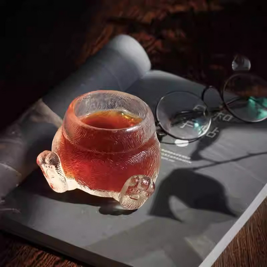 A cozy lifestyle scene featuring a cat paw glass teacup on an open book next to vintage glasses, glowing with warm tea in a soft evening light.
