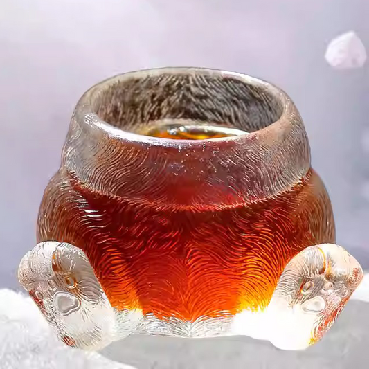 A whimsical handcrafted glass teacup shaped like a cat's paws and fluffy back, featuring detailed fur texture and glowing with warm amber tea.