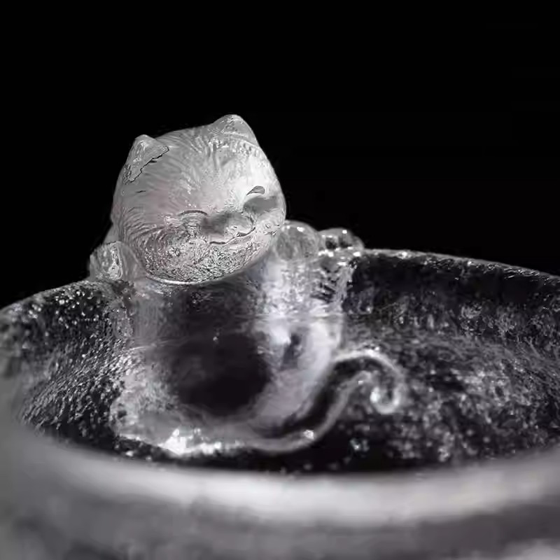 An adorable handcrafted glass teacup featuring a smiling cat sculpture on the rim, with a unique hammered ice-crackle texture.