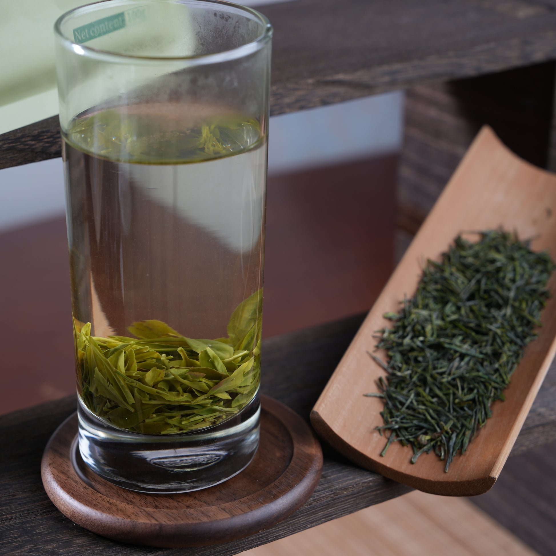 A tall glass of freshly brewed Enshi Yulu green tea showing leaves elegantly unrolling, displayed next to dry pine-needle shaped loose leaves on a bamboo scoop.