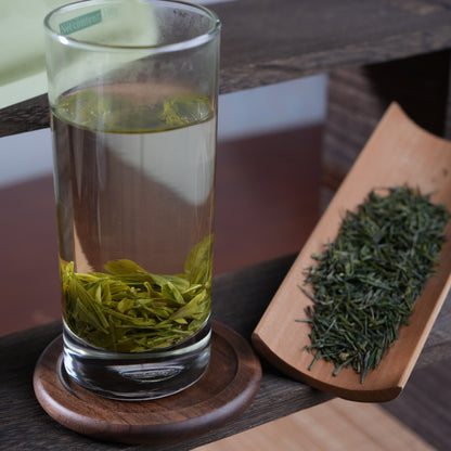 A tall glass of freshly brewed Enshi Yulu green tea showing leaves elegantly unrolling, displayed next to dry pine-needle shaped loose leaves on a bamboo scoop.