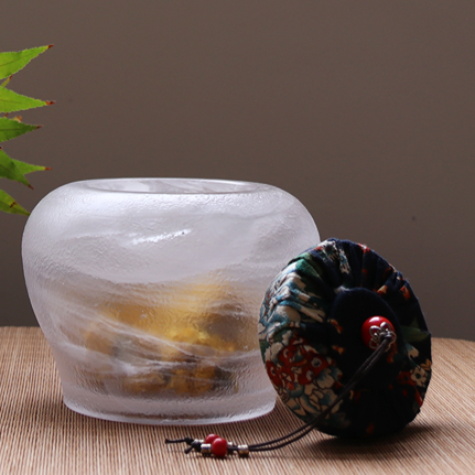 An elegant frosted glass tea caddy with an ice-crackle texture, featuring a traditional floral fabric lid with a red bead pull for airtight storage.