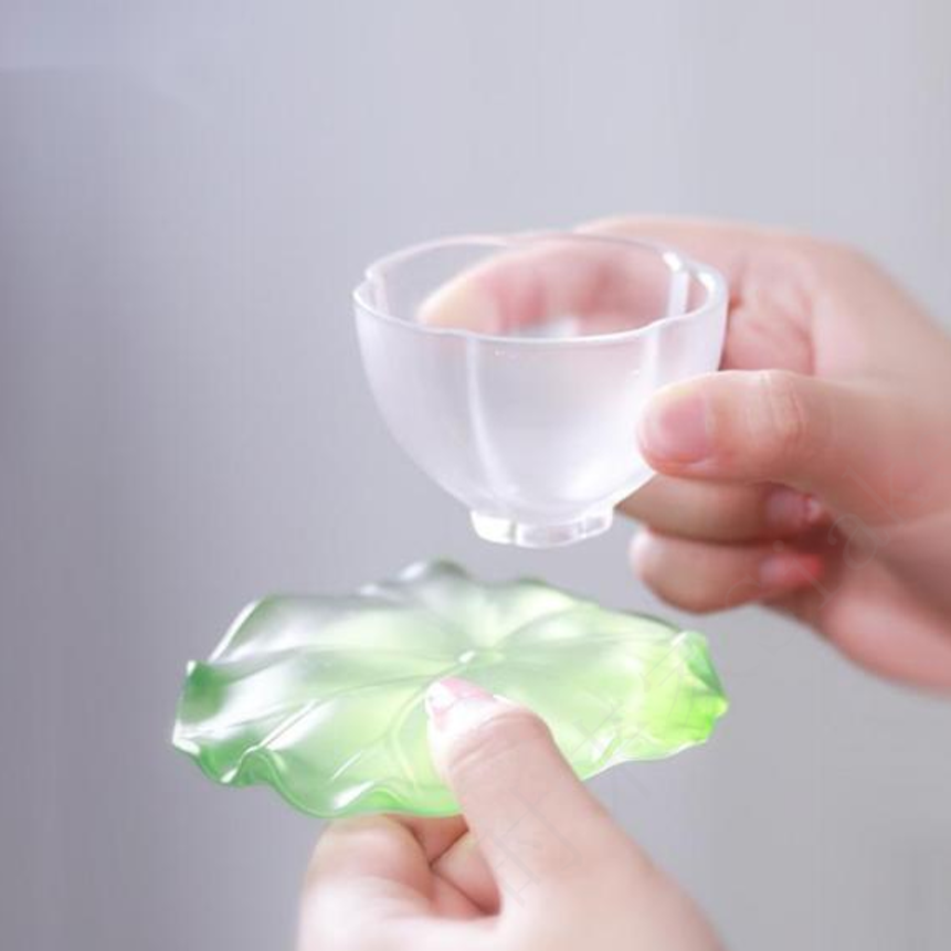 A beautiful Zen tea set featuring a frosted petal-shaped glass cup and a translucent green lotus leaf saucer, held elegantly to showcase a refreshing spring aesthetic.