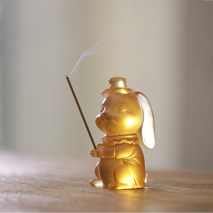 A whimsical yellow translucent rabbit figurine wearing a top hat, functioning as an incense holder with wisps of smoke, set in a serene tea-room environment.