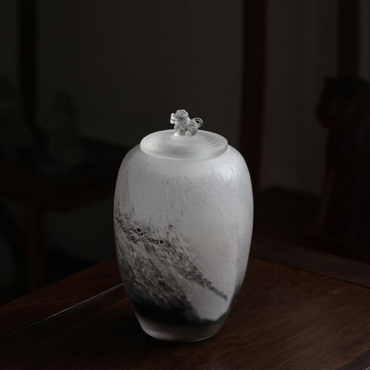 An artistic frosted glass tea caddy featuring a dramatic black ink-wash design and a transparent guardian lion sculpture on the lid, set against a dark wooden background.