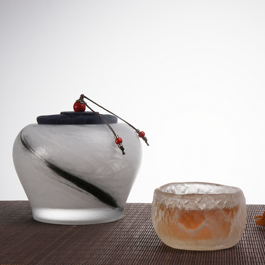An artistic tea set featuring a frosted glass tea caddy with a black ink-wash stroke and a matching textured ice-crackle teacup on a bamboo mat.