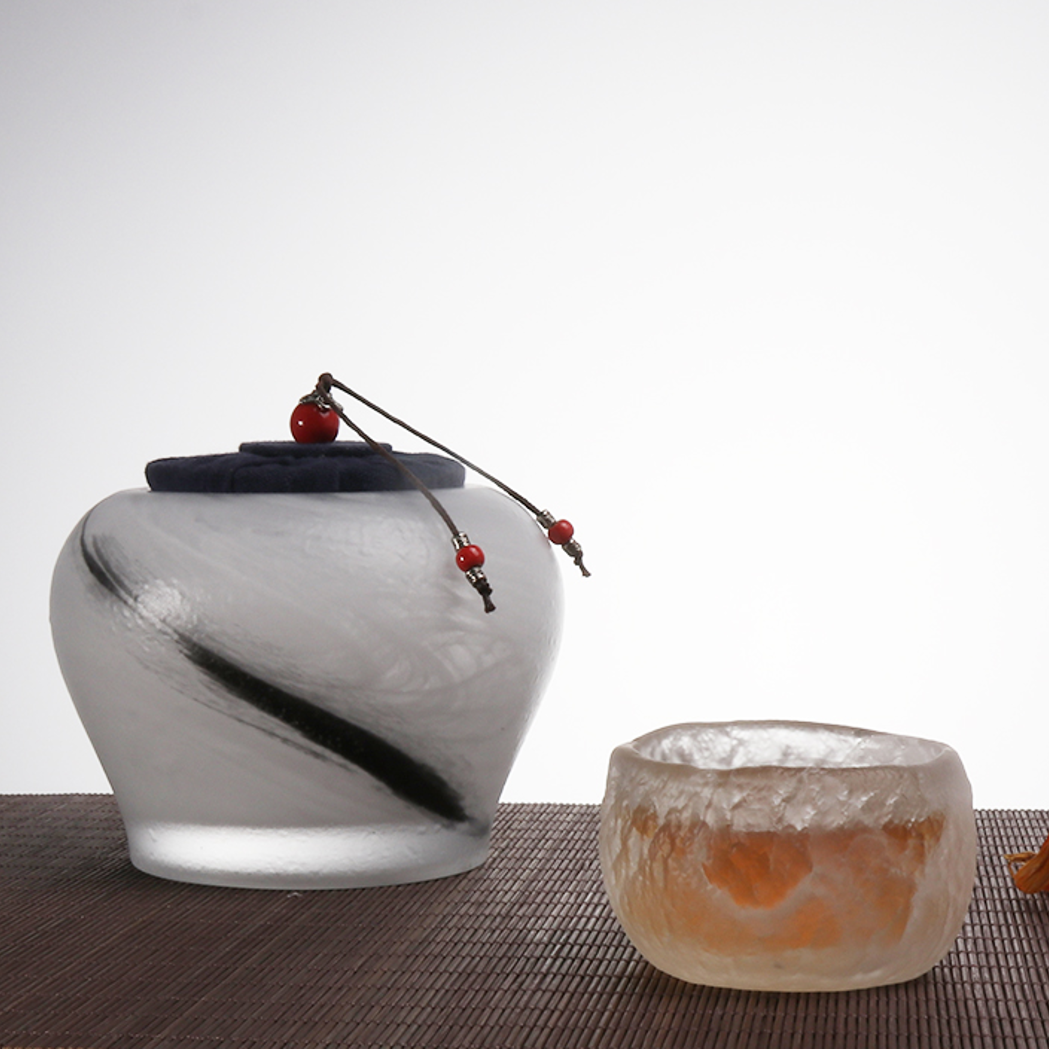 An artistic tea set featuring a frosted glass tea caddy with a black ink-wash stroke and a matching textured ice-crackle teacup on a bamboo mat.