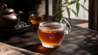 Handcrafted Menhai Imperial Cat liuli tea cup filled with amber tea, featuring a clear cat-shaped handle in a traditional Chinese tea setting.
