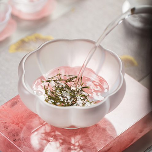 Fresh water being poured into a frosted petal-shaped glass cup filled with premium needle-shaped green tea leaves, showcasing a delicate brewing ritual.