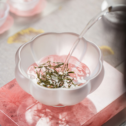 Fresh water being poured into a frosted petal-shaped glass cup filled with premium needle-shaped green tea leaves, showcasing a delicate brewing ritual.