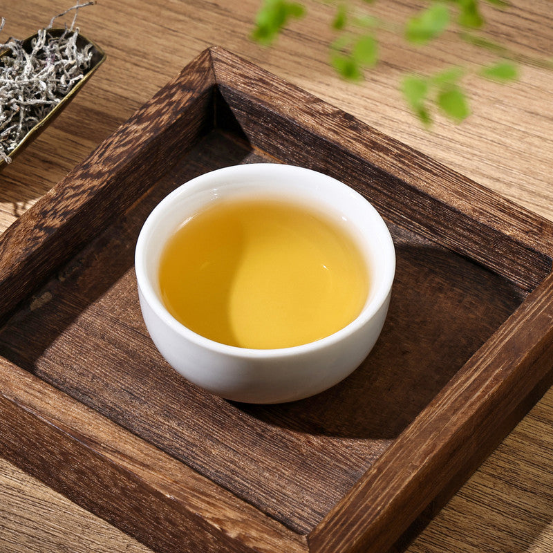 A white ceramic cup of freshly brewed golden Vine Tea by SeTeng Tea, presented in a rustic square wooden tray with dry leaves in the background.